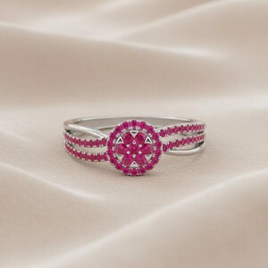 Könnte beinhalten: Ein silberner Ring mit einem zentralen Cluster aus rosa Edelsteinen, umgeben von einem Kranz aus kleineren rosa Steinen. Der Ring hat drei Reihen rosa Edelsteine auf jeder Seite des zentralen Clusters, was einen geschichteten Effekt erzeugt.