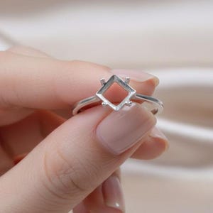 Könnte beinhalten: Ein silberner Ring mit rautenförmiger Fassung. Der Ring wird von einer Person gehalten. Die Fassung ist offen und für einen Edelstein bestimmt. Der Ring hat ein dünnes, schlichtes Band.