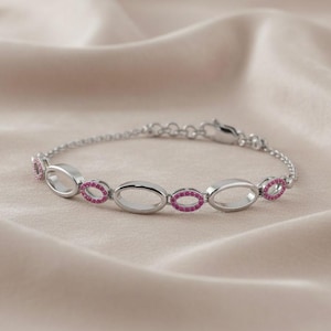 Könnte beinhalten: Ein silbernes Armband mit ovalen Gliedern, die jeweils mit einem Ring aus kleinen, rosa Edelsteinen besetzt sind. Das Armband liegt auf einer weichen, beigefarbenen Oberfläche. Der Verschluss ist sichtbar.