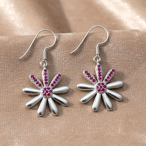 Könnte beinhalten: Ein Paar silberner Ohrringe in Form einer Gänseblümchenblüte mit Hakenverschluss. Jeder Ohrring hat ein Blumendesign mit silbernen Blütenblättern und einem Zentrum aus kleinen, runden, rosa Edelsteinen. Die Ohrringe werden auf einer beigen Oberfläche präsentiert.