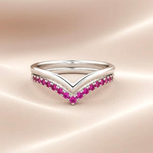 Könnte beinhalten: Ein Set aus zwei silbernen Ringen. Ein Ring ist ein schlichtes Band, der andere ein V-förmiger Ring mit einer Reihe kleiner, leuchtend rosa Edelsteine. Die Ringe sind vor einem weichen, neutralen Hintergrund platziert.