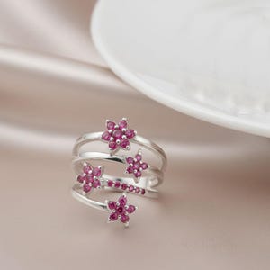Könnte beinhalten: Ein silberner Ring mit floralem Design. Der Ring verfügt über mehrere Bänder, die mit kleinen, leuchtend rosa blütenförmigen Edelsteinen verziert sind. Die Blüten sind zusammengefasst und bilden ein zartes und auffälliges Schmuckstück.