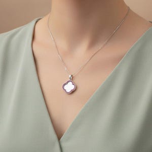 Könnte beinhalten: Eine silberne Halskette mit einem kleeblattförmigen Anhänger. Der Anhänger hat eine weiße Mitte und einen Rand aus kleinen, rosa Edelsteinen. Die Halskette wird auf einem hellgrünen Oberteil getragen.