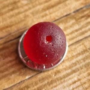 May include: A round, deep red bead with a small hole, resting on a silver coin. The bead has a textured surface and is set against a wooden background. This bead is suitable for jewelry making.