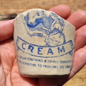 Op de afbeelding: Fragment van een roomcontainer met een blauw-wit ontwerp. Het woord "CREAM" is gedrukt in een banner, met een illustratie van een koe en een vrouw. De tekst "Cream contains a small quantity of preservative to prolong its smell" is ook zichtbaar.