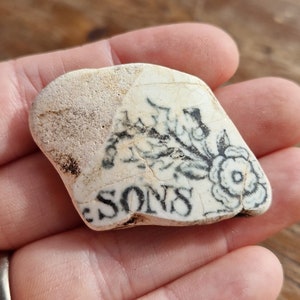May include: A white ceramic shard with black ink markings. The shard features a floral design and the word "SONS".