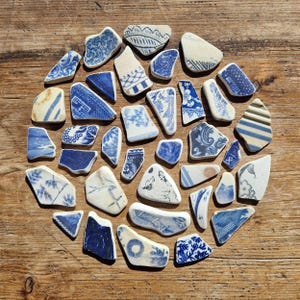 Puede incluir: Una colección de fragmentos de cerámica azul y blanca dispuestos en un patrón circular sobre una superficie de madera. Los fragmentos presentan varios diseños, incluyendo motivos florales y formas geométricas. La paleta de colores es principalmente azul y blanco.