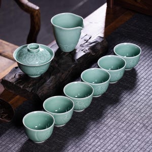 May include: A celadon green tea set featuring a lidded bowl, a pitcher, and six small cups. The porcelain pieces have a floral pattern and are arranged on a dark wooden surface, suggesting a traditional tea ceremony.
