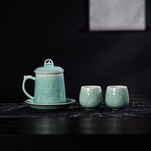 Puede incluir: Un juego de té verde celadón con una taza con tapa y asa, un platillo y dos tazas pequeñas. El juego tiene un diseño floral tallado. El fondo es una superficie oscura con una pintura borrosa.