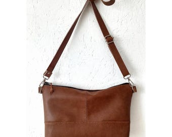 May include: A medium brown crossbody bag featuring a rectangular body with a horizontal seam across the lower section. The bag includes a long, adjustable shoulder strap with silver-toned metal hardware and a top zipper closure with a small pull tab.