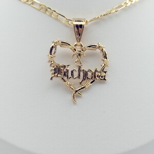 May include: A gold-coloured necklace with a heart-shaped pendant. The pendant features the word "Bichota" in a stylised font, surrounded by a barbed wire design. The necklace has a chain with a rectangular link pattern.