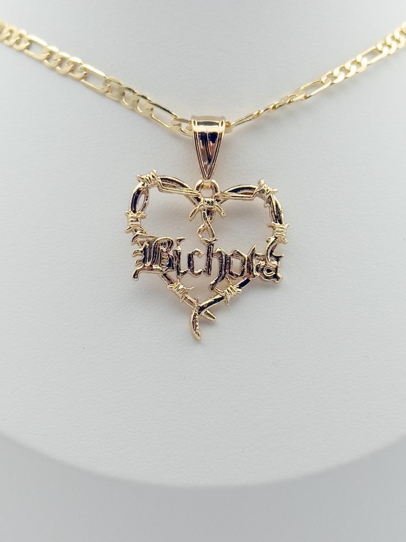 May include: A gold-coloured necklace with a heart-shaped pendant. The pendant features the word "Bichota" in a stylised font, surrounded by a barbed wire design. The necklace has a chain with a rectangular link pattern.