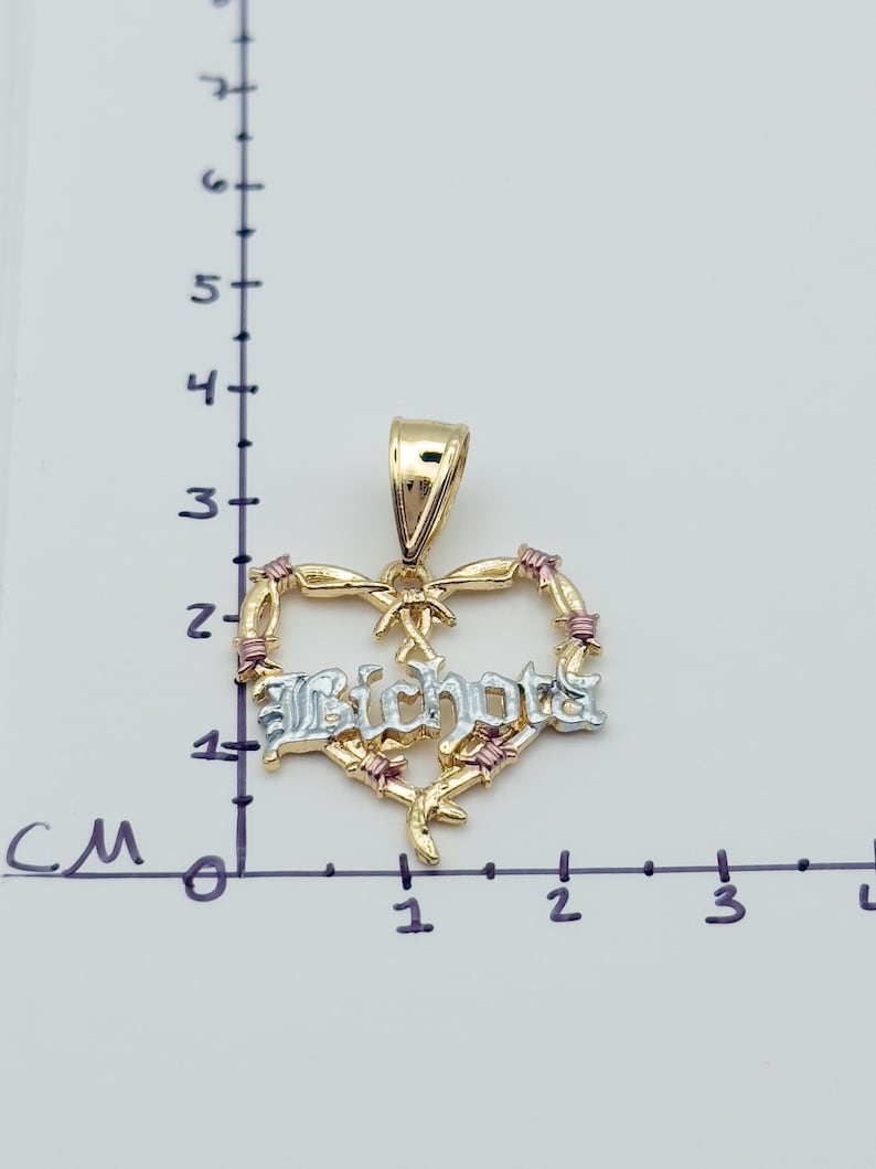 May include: A gold heart pendant with the word "Bichota" in silver lettering. The heart is decorated with pink accents and a bow. The pendant is approximately 1.5 cm wide.