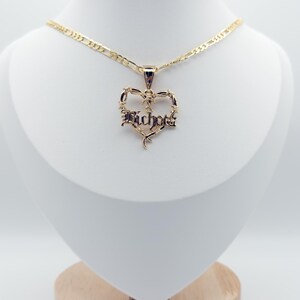 May include: A gold necklace with a heart pendant. The pendant has the word "Bichota" in a gothic font, framed by a barbed wire design. The necklace is displayed on a white mannequin.
