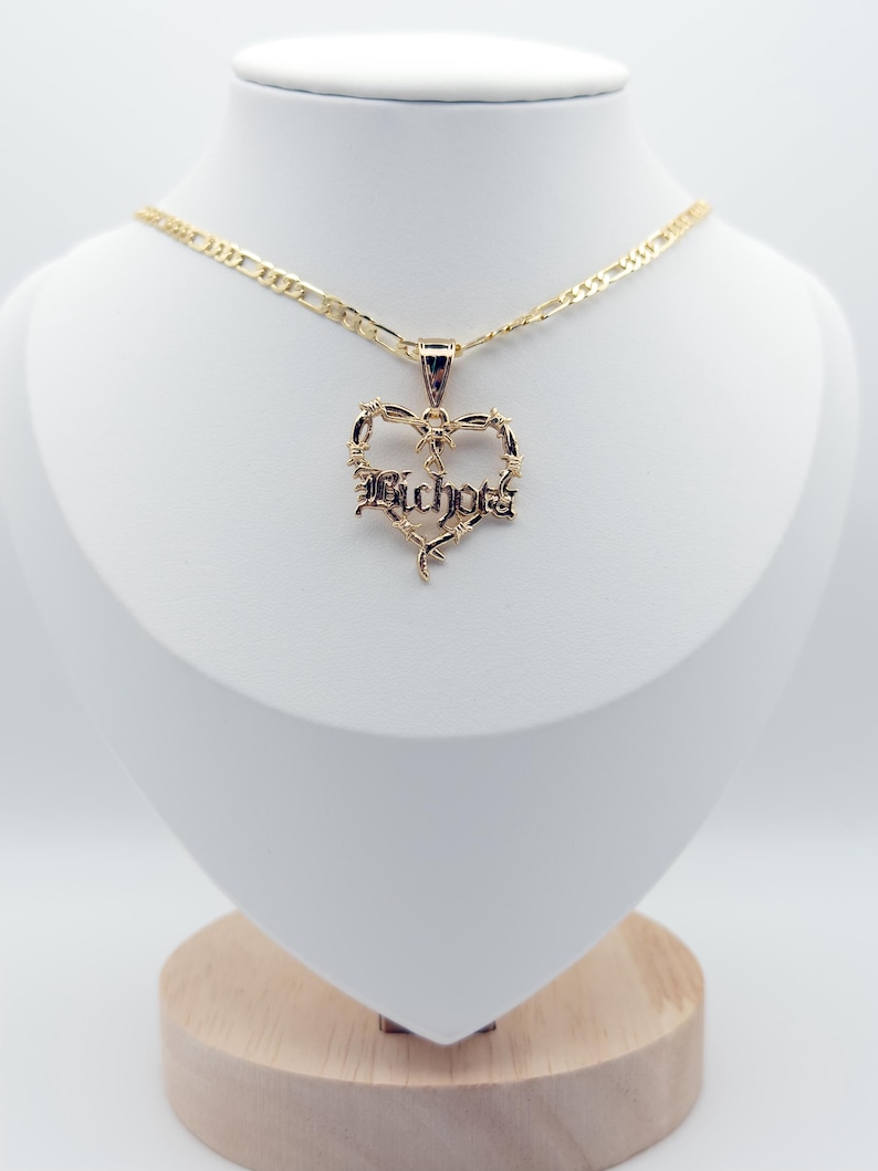 May include: A gold necklace with a heart pendant. The pendant has the word "Bichota" in a gothic font, framed by a barbed wire design. The necklace is displayed on a white mannequin.