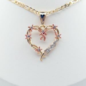 May include: A gold-coloured heart pendant necklace. The heart features small pink flowers and a ribbon design. The word "Isibola" is written on the heart. The necklace chain is a gold figaro chain.