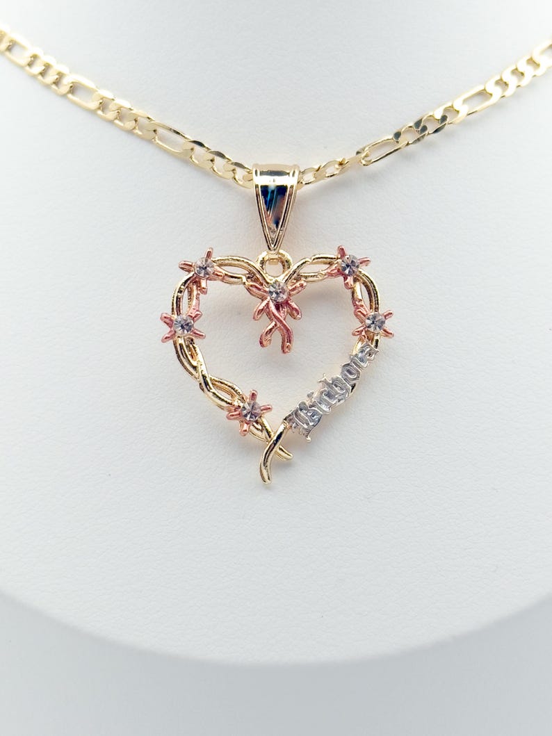 May include: A gold-coloured heart pendant necklace. The heart features small pink flowers and a ribbon design. The word "Isibola" is written on the heart. The necklace chain is a gold figaro chain.