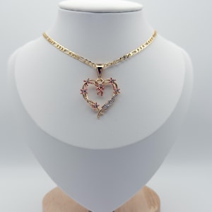 May include: A gold necklace with a heart pendant. The pendant is a heart shape decorated with small pink and clear gemstones. The necklace is displayed on a white mannequin against a white backdrop.