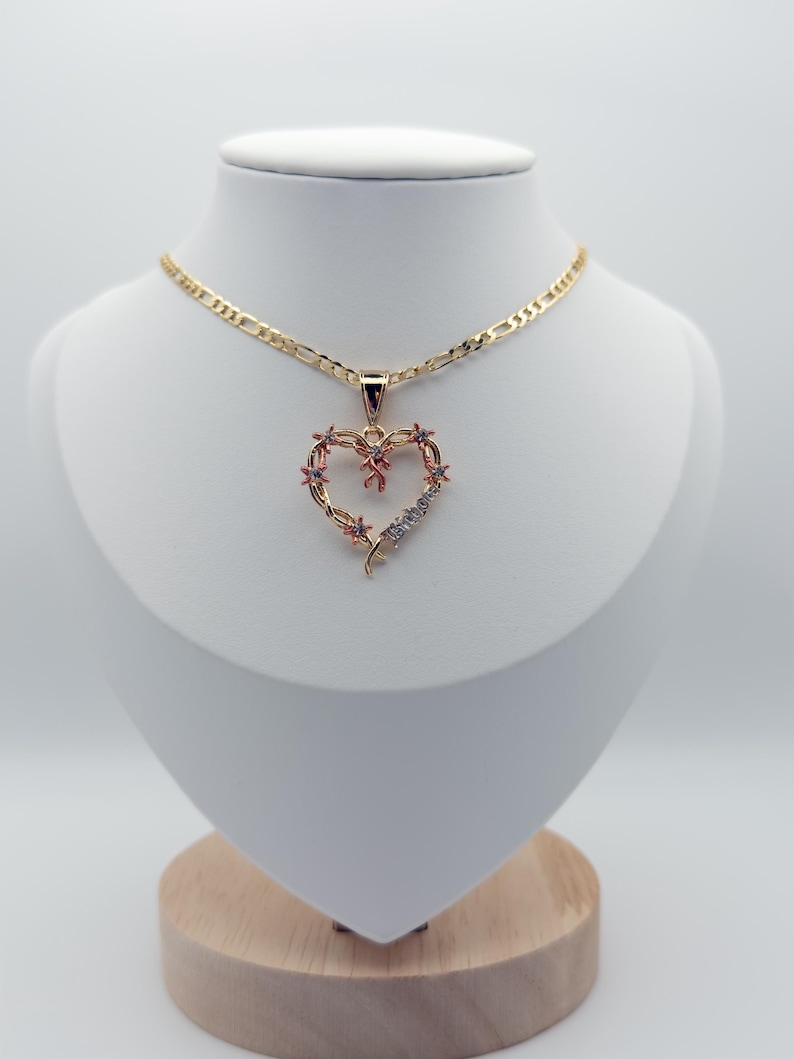 May include: A gold necklace with a heart pendant. The pendant is a heart shape decorated with small pink and clear gemstones. The necklace is displayed on a white mannequin against a white backdrop.