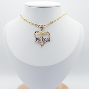 May include: A gold necklace with a heart-shaped pendant. The pendant has the word "Bichota" in a gothic font, with pink and blue detailing. The necklace is displayed on a white mannequin.