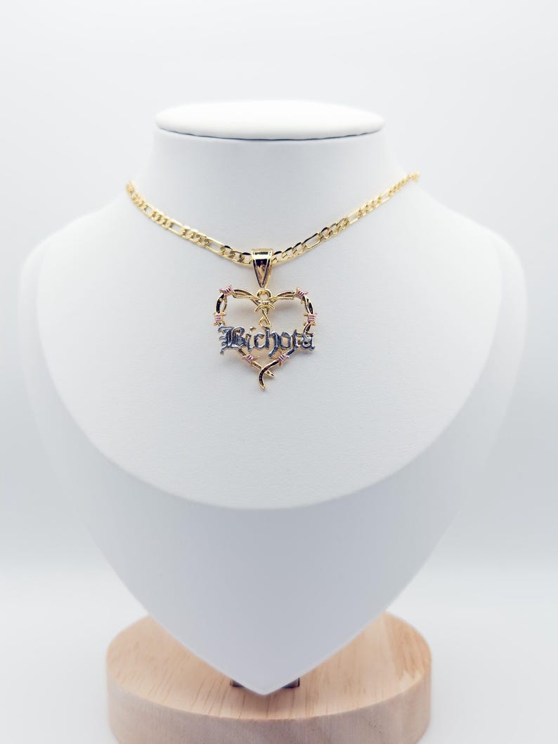 May include: A gold necklace with a heart-shaped pendant. The pendant has the word "Bichota" in a gothic font, with pink and blue detailing. The necklace is displayed on a white mannequin.