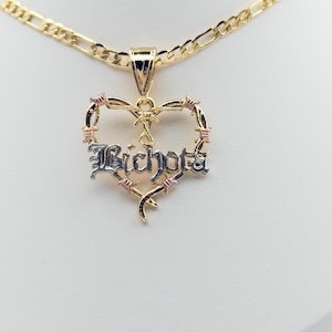 May include: A gold necklace with a heart-shaped pendant. The pendant features the word "Bichota" in a silver font, framed by a barbed wire design with pink accents. The necklace has a gold chain.
