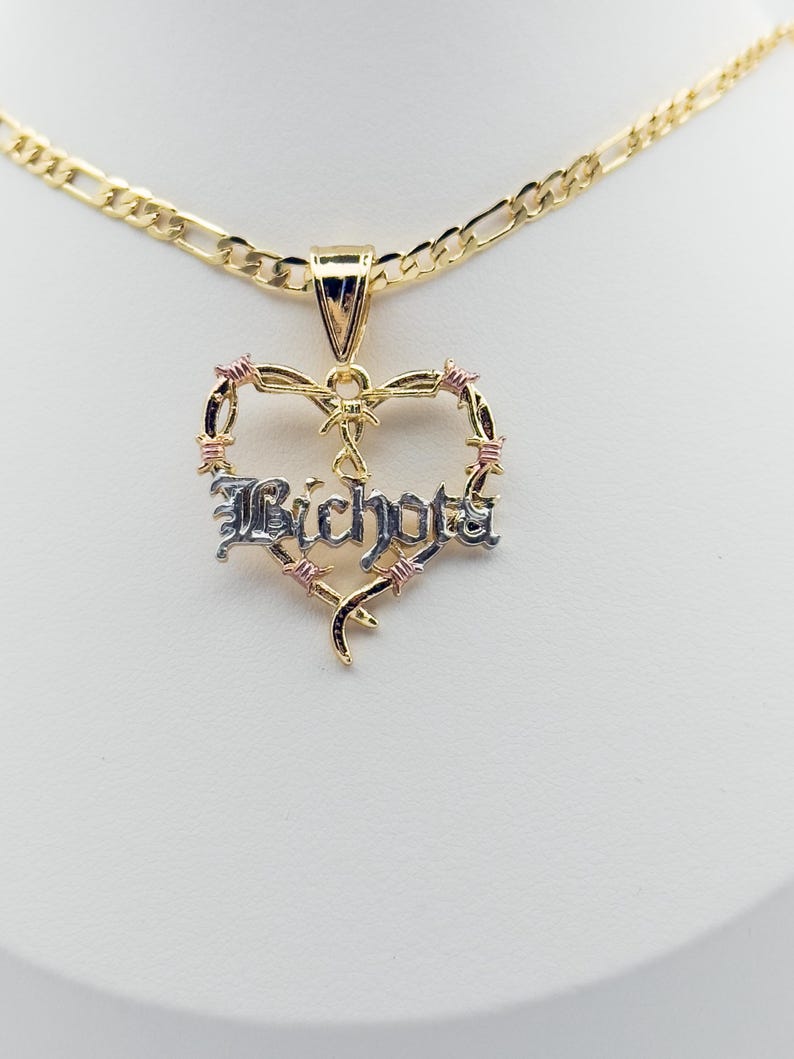 May include: A gold necklace with a heart-shaped pendant. The pendant features the word "Bichota" in a silver font, framed by a barbed wire design with pink accents. The necklace has a gold chain.