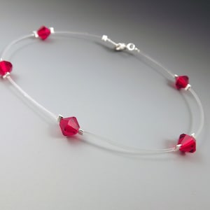 May include: A delicate necklace featuring five faceted, ruby-red crystals strung on a clear, nearly invisible cord. The crystals are evenly spaced and secured with small silver-colored metal accents. The necklace has a simple clasp.