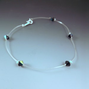 May include: A delicate necklace featuring small, faceted, dark-colored beads spaced along a thin, clear cord. The necklace has a silver-colored clasp. The beads have a subtle iridescent quality.