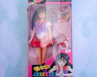 May include: A Sailor Moon doll in its original packaging. The doll is wearing a red, pink, and white outfit with a purple bow. The box features the Sailor Moon logo and character illustrations.