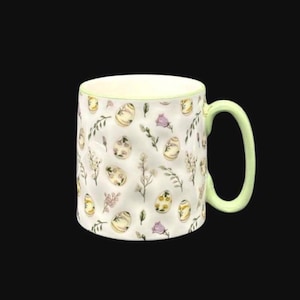 May include: White ceramic mug with a light green handle and rim. The mug is decorated with a pattern of Easter eggs, flowers, and leaves in shades of yellow, green, and purple. The mug is approximately 4 inches tall.