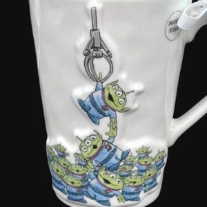 Puede incluir: Taza de cer&aacute;mica blanca con un dise&ntilde;o de extraterrestre de dibujos animados. La taza representa extraterrestres verdes con uniformes azules, uno de los cuales es levantado por una garra. La parte inferior de la taza tiene un patr&oacute;n repetido de los mismos extraterrestres.