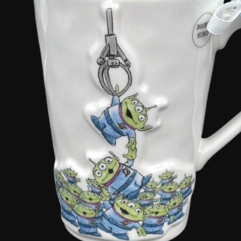 Puede incluir: Taza de cer&aacute;mica blanca con un dise&ntilde;o de extraterrestre de dibujos animados. La taza representa extraterrestres verdes con uniformes azules, uno de los cuales es levantado por una garra. La parte inferior de la taza tiene un patr&oacute;n repetido de los mismos extraterrestres.