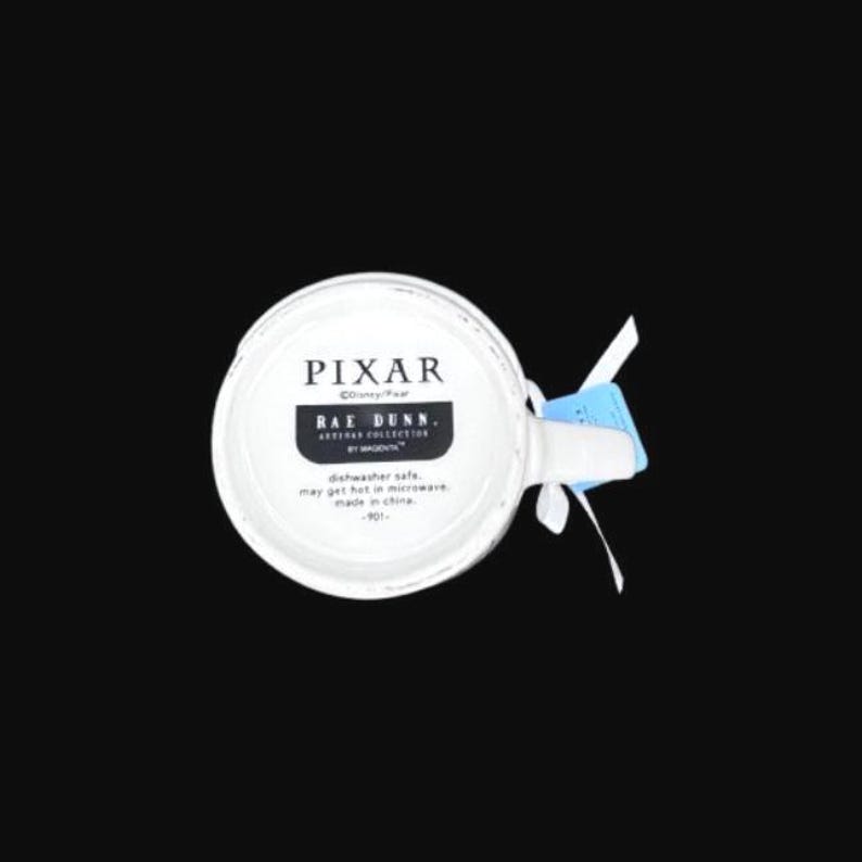 Puede incluir: Taza de cer&aacute;mica blanca con el texto "PIXAR" y "RAE DUNN" en letras negras. La taza tiene un asa y una etiqueta azul adjunta. La taza es apta para lavavajillas y microondas.