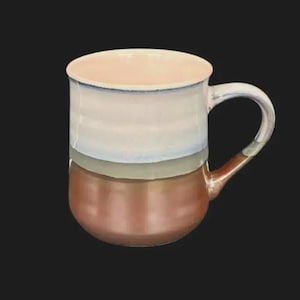 May include: A ceramic mug with a rounded base and a handle. The mug features a colour-blocked design with a light pink interior, white and blue upper section, a grey band, and a copper-coloured lower section.