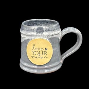 May include: Gray ceramic mug with a handle and a gold circular label. The label features the words "love your melon" in a handwritten style. The mug has a textured, glazed finish and a dark background.