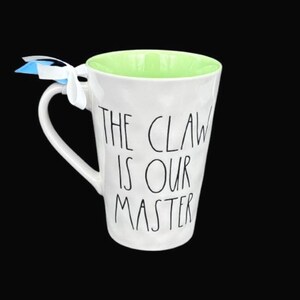 Puede incluir: Taza de cer&aacute;mica blanca con interior verde y el texto "THE CLAW IS OUR MASTER" en negro. La taza tiene un asa curva y est&aacute; atada con una cinta blanca y una etiqueta azul y blanca. La taza est&aacute; sobre un fondo negro.
