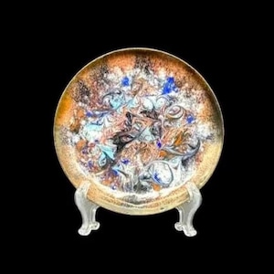 May include: Decorative plate with a unique abstract design in shades of brown, blue, and white. The plate has a gold-colored rim and is displayed on a clear, ornate stand. The plate's design features swirling patterns.