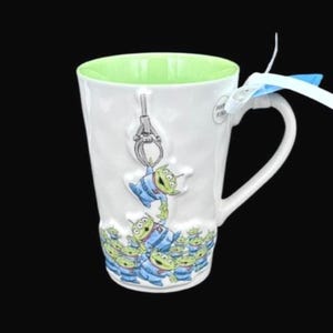 Puede incluir: Taza de cer&aacute;mica blanca con interior verde y asa curva. La taza est&aacute; decorada con un dise&ntilde;o de extraterrestres verdes con uniformes azules, con un gr&aacute;fico de m&aacute;quina de garra. Una cinta azul y blanca est&aacute; atada al asa.