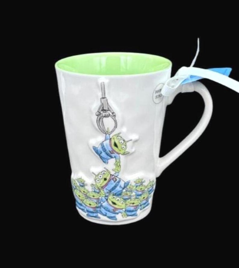 Puede incluir: Taza de cer&aacute;mica blanca con interior verde y asa curva. La taza est&aacute; decorada con un dise&ntilde;o de extraterrestres verdes con uniformes azules, con un gr&aacute;fico de m&aacute;quina de garra. Una cinta azul y blanca est&aacute; atada al asa.