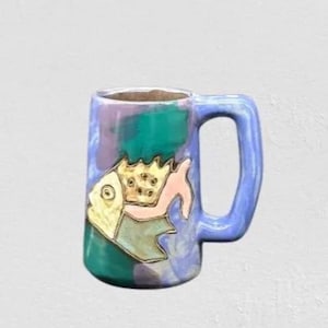 May include: A ceramic mug with a blue handle and body, featuring a hand-painted fish design. The fish has gold, pink, and teal accents. The mug's body has a mottled pattern of teal, purple, and blue. The interior is a dark brown color.