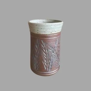 Peut inclure: Un vase en céramique fait à la main de forme cylindrique. Le vase présente un extérieur marron texturé avec un motif de tiges de blé en relief. Le bord est de couleur blanc cassé moucheté. Le vase mesure environ 15 cm de haut.