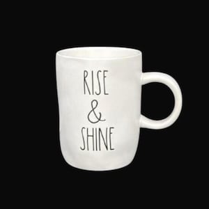 Puede incluir: Taza de cerámica blanca con asa curva. La taza presenta las palabras "RISE & SHINE" en negro, texto alineado verticalmente. La taza está sobre un fondo negro.