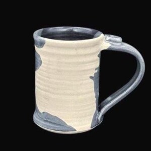 May include: A cylindrical ceramic mug featuring a cream-colored body with horizontal ribbed texture and irregular dark gray patches. The mug has a smooth, curved handle in a matching dark gray glaze, and the interior is finished in a dark gray tone.