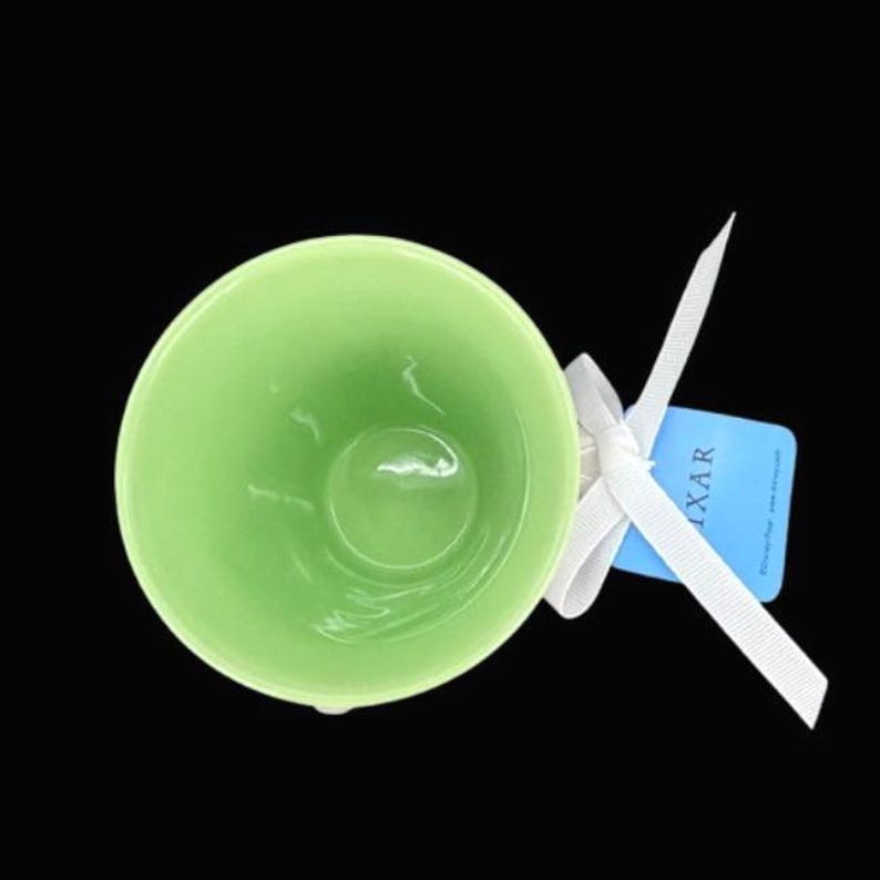 Puede incluir: Una taza de cer&aacute;mica verde claro con forma redondeada. Una cinta blanca est&aacute; atada alrededor de la taza, y una etiqueta azul con la palabra "IXAR" est&aacute; adjunta. La taza est&aacute; sobre un fondo negro.