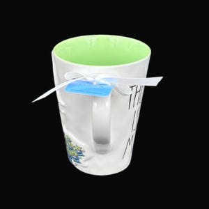 Puede incluir: Taza de cer&aacute;mica blanca con interior verde y asa. La taza tiene el texto "THE IS ME" en letras negras. Una etiqueta azul est&aacute; sujeta con una cinta blanca. Un peque&ntilde;o gr&aacute;fico de extraterrestres verdes y azules est&aacute; en el lateral.