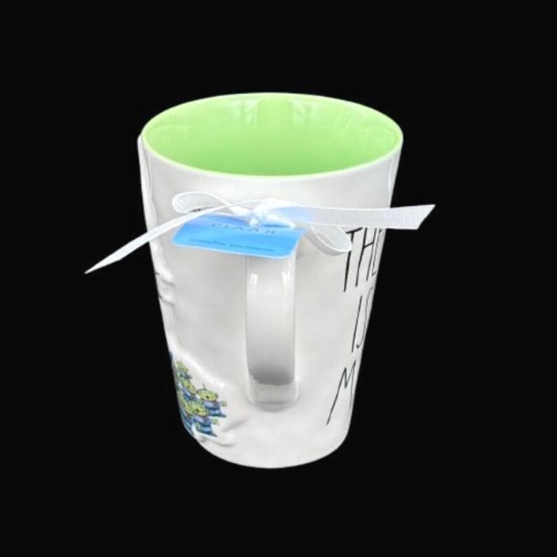 Puede incluir: Taza de cer&aacute;mica blanca con interior verde y asa. La taza tiene el texto "THE IS ME" en letras negras. Una etiqueta azul est&aacute; sujeta con una cinta blanca. Un peque&ntilde;o gr&aacute;fico de extraterrestres verdes y azules est&aacute; en el lateral.