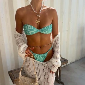 Peut inclure: Ensemble de bikini à motifs turquoise et blancs. Le modèle porte un cache-maillot blanc en tricot ajouré à manches longues et une ceinture chaîne dorée. Un collier de perles et d'étoiles de mer et un petit sac à main beige à chaîne dorée complètent le look.