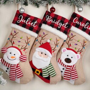 May include: Three Christmas stockings with personalized names: Juliet, Bridget, and Caroline. Each stocking has a red and black plaid cuff, a beige body with embroidered antlers and lights, and a character face: snowman, Santa, and a polar bear.