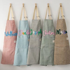 May include: Five striped aprons in shades of red, blue, grey, green, and brown hang on a white wall. Each apron features a name in colourful script: Wallace, Claudia, Joseph, Victor, and Maria. The aprons have light-coloured straps.